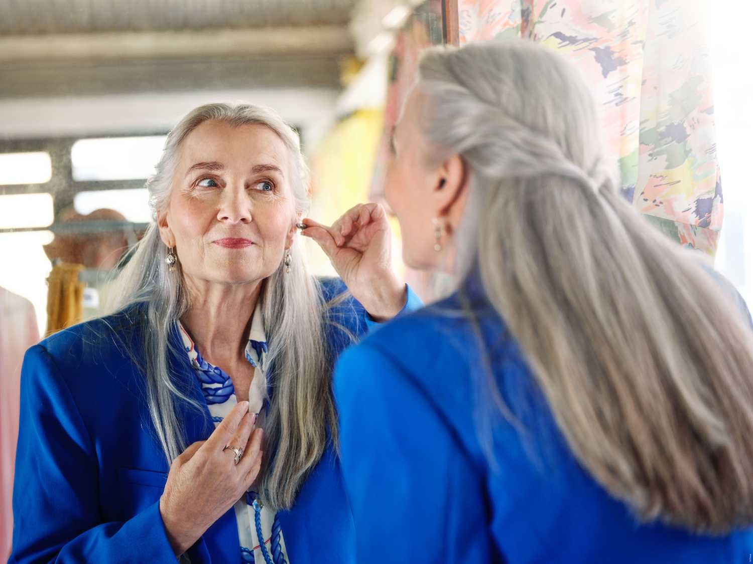 Frau mit grauen Haaren setzt ein Hörgerät vor einem Spiegel ein