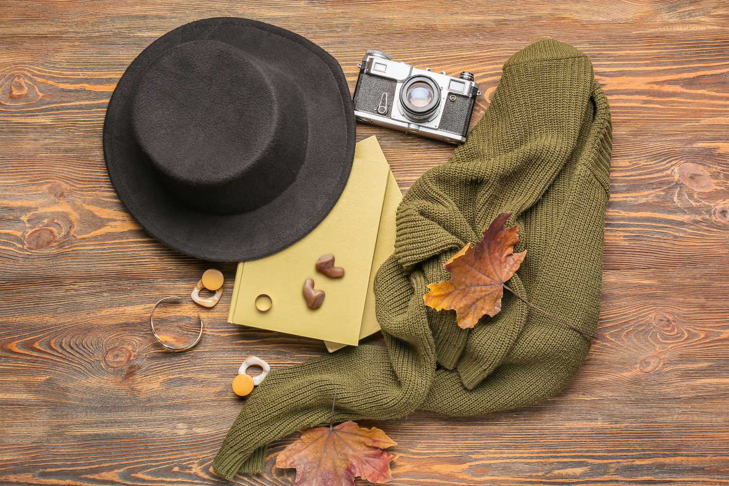 Auf einem Tisch mit Holzmaserung liegt ein Paar Hörgeräte auf einem gelben Brief samt Umschlag zwischen verschiedenen Gegenständen, wie einem olivgrünen Pullover, Herbstblättern, einem schwarzen Hut und einer antiken Kamera.